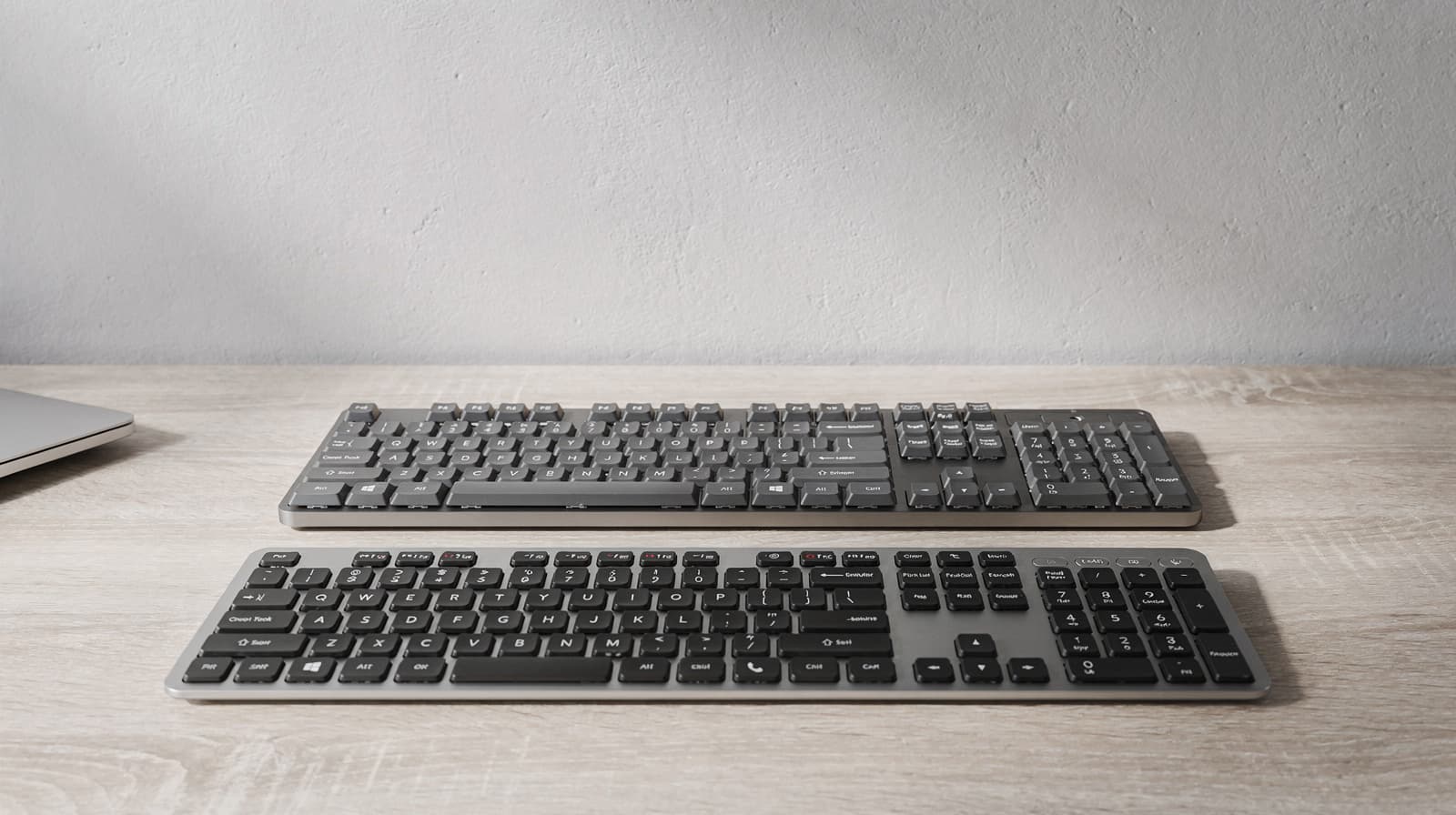 Two graphite office keyboards with flat and raised keys on a light wood desk